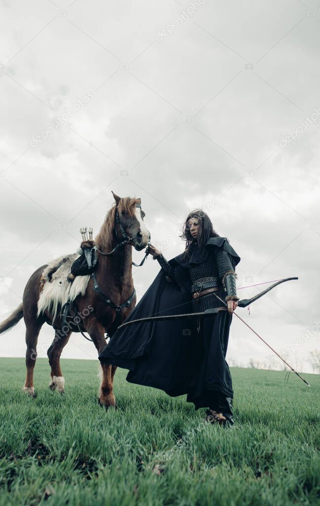 Woman in chain mail in image of medieval warrior stands with bow in her hand near horse in field.