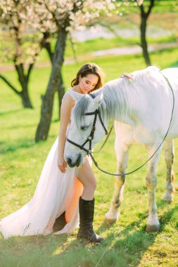 Uzun beyaz elbiseli gelin beyaz atın yanında duruyor ve onu bahar bahçesinde çiçek açan kayısı ağaçlarının arka planında kucaklıyor..