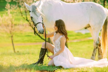 Uzun beyaz elbiseli gelin beyaz atın yanında oturuyor ve onu çayırda tutuyor..