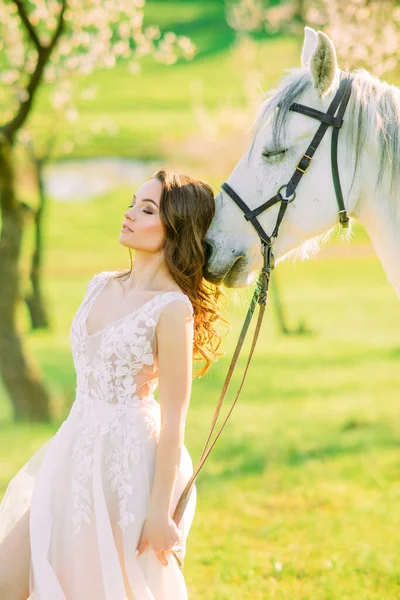 Bahar bahçesinde beyaz at başına yaslanmış uzun beyaz elbiseli gelin çiçek açan kayısı ağaçlarının arka planında.