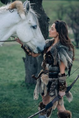 Kürk giymiş bir kadın, atının yanındaki ormanda elinde yayla antik savaşçı Amazon suretinde duruyor..