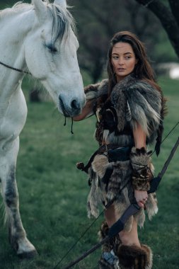 Woman stands in fur clothes in image of ancient warrior amazon with bow in her hands in forest near her horse. clipart
