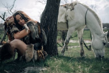 Kadın ormanda çimlerin üzerinde oturur ve eski savaşçı Amazon suretinde elinde kılıçla otlayan atın arka planında dinlenir..
