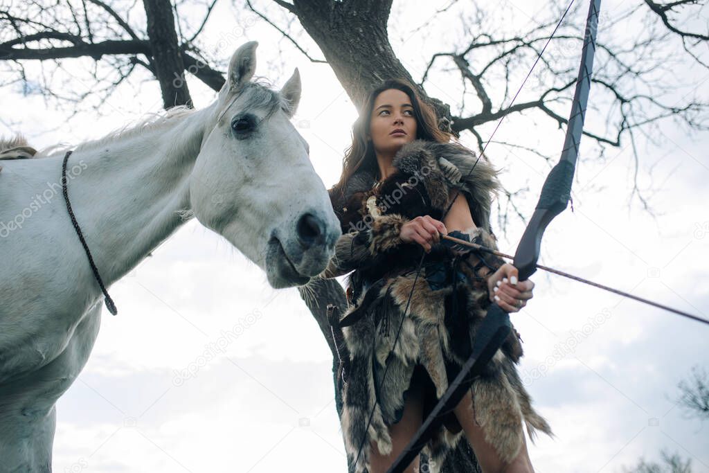 Woman in image of ancient warrior amazon shoots a bow and aims at target near her horse among forest.