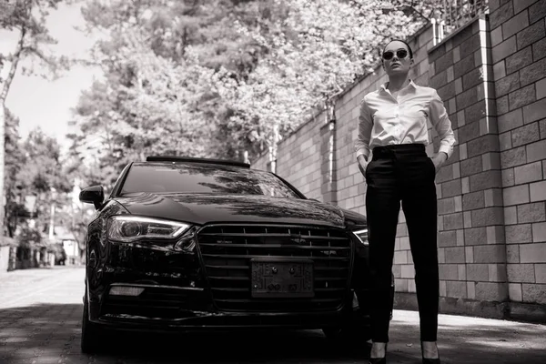 戴着太阳镜的年轻漂亮的女人站在城市街道上的一辆黑色现代汽车旁边 黑白图像 — 图库照片