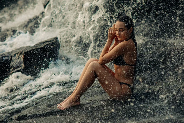 美しいですセクシー女性は滝の流れの下ビキニに座って 目を閉じて落下水を楽しむ — ストック写真