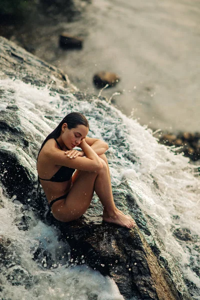 美しいですセクシー女性は滝の流れの下ビキニに座って 落下水を楽しんでいます — ストック写真