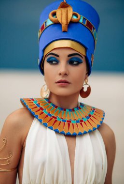 Portrait of young woman in image of ancient egyptian queen Nefertiti against background of sandy desert and blue sky.