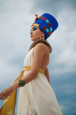 Young woman in image of ancient egyptian queen Nefertiti walks against background of blue sky.