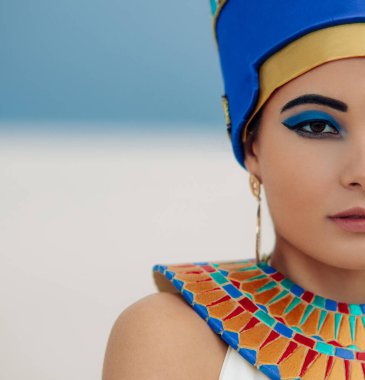 Half-portrait of young woman in image of ancient egyptian queen Nefertiti against background of sandy desert and blue sky.