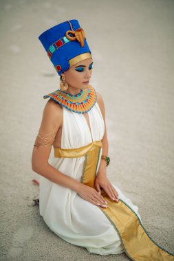 Young woman in image of ancient egyptian queen Nefertiti sits in sandy desert.