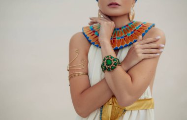Jewelry and dress of young woman in image of ancient egyptian queen Nefertiti against background of sandy desert. Closeup.