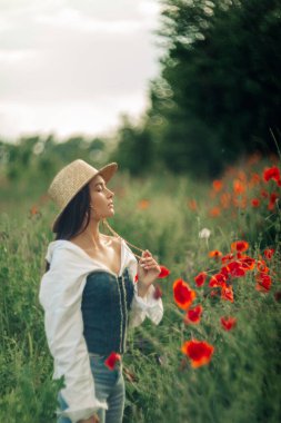 Hasır şapkalı genç kadın çayırların arasında çiçek açan gelinciklerle eğleniyor..