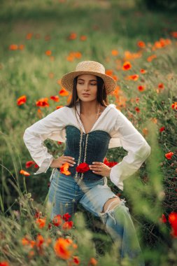 Hasır şapkalı genç kadın çayırların arasında çiçek açan gelinciklerle eğleniyor..