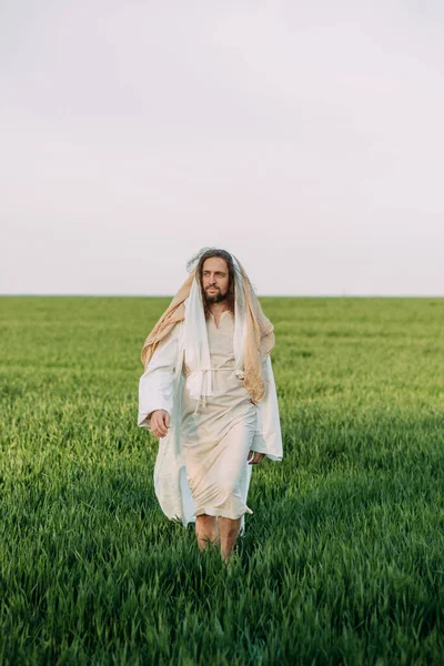 Jesus Christ walking in meadow clothed in his traditional white robe against sky background.