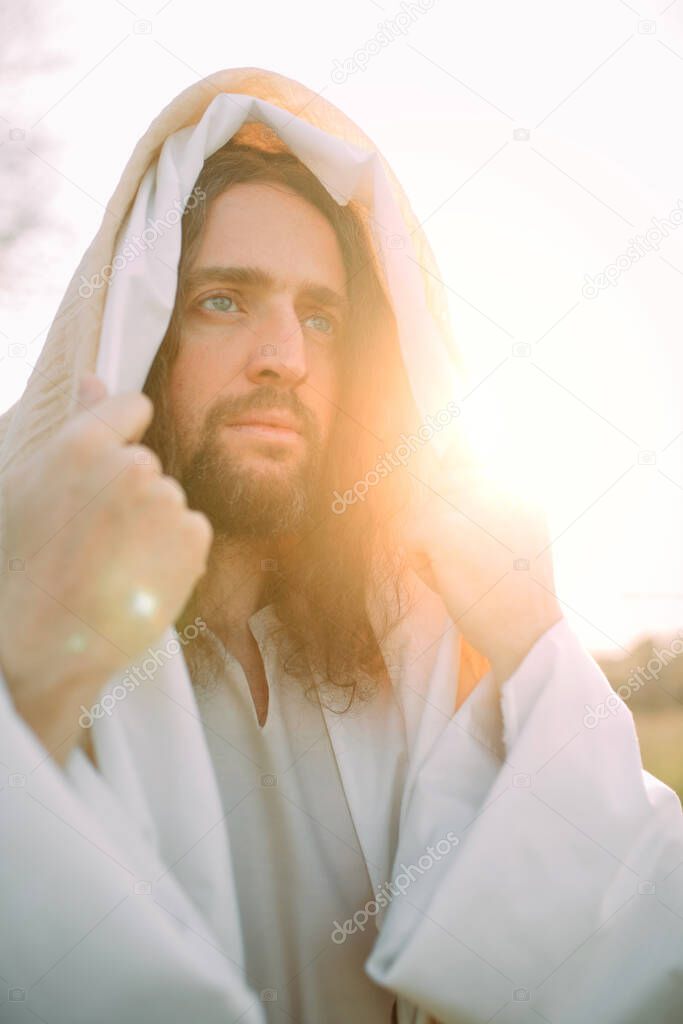 Portrait of Jesus Christ standing in meadow clothed in his traditional white robe against sunset background.