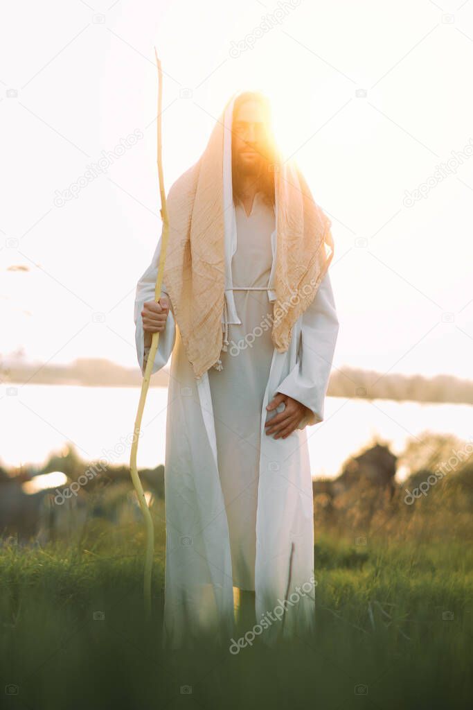 Jesus Christ with wooden staff stands in meadow clothed in his traditional white robe against river and sunset background.