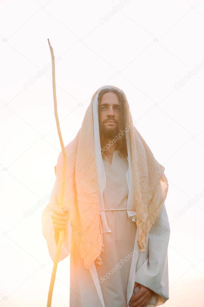 Jesus Christ with wooden staff stands in his traditional white robe against sky and sunset background.
