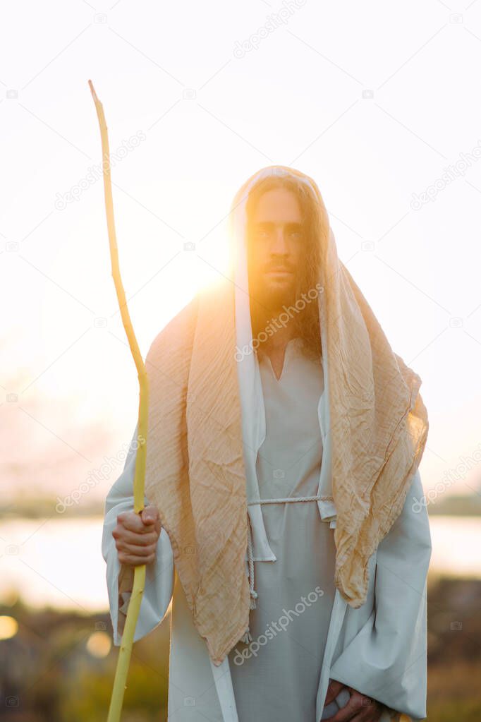 Jesus Christ with wooden staff stands in meadow clothed in his traditional white robe against river and sunset background.