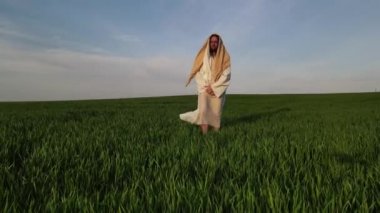 İsa, geleneksel beyaz cübbesiyle gökyüzü arka planında çayırlarda yürür..