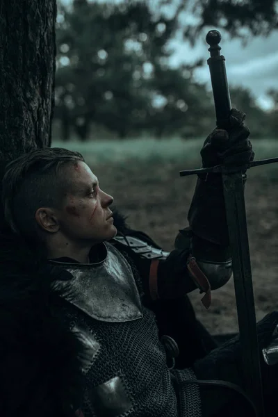 Medieval tired warrior knight in chain mail armour sits and rests among forest under tree with sword in his hand.