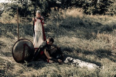 Eski Spartalı savaşçı savaş elbisesi ve kadın okçu köpekle birlikte kalkanın ve mızrağın yanındaki çayırda dinleniyor..