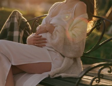 Güzel hamile bir kadın, altın saat günbatımında parktaki bankta dinlenirken bebek çarpmasına hafifçe dokunuyor. Annelik beklentisi aşkını kavra.