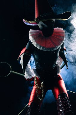 Karanlık soytarı ya da palyaço figürünün dramatik yakın plan Cadılar Bayramı karakteri canlı kırmızı ve siyah bir kostüm içinde, tiyatro ışığıyla aydınlatılmış ve dumanla çevrili..