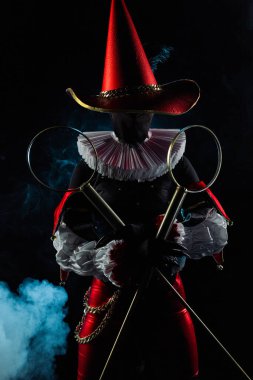 Çarpıcı bir soytarı ya da palyaço figürü canlı kırmızı ve siyah bir kostüm içinde, büyük bir konik şapka ve yaka, siyah dumanlı arka planda baloncuk asalar tutuyor. Cadılar Bayramı karakter teması.