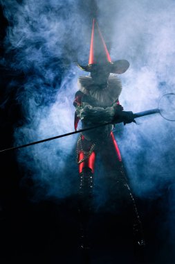 Kara cadılar bayramı teması için siyah ve kırmızı kostümlü soytarı ya da soytarı karakteri dramatik bir mavi duman ve siyah arka planda ışıklandırma ile sahnede duruyor..