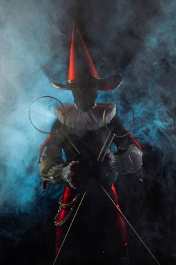 Çarpıcı bir soytarı ya da palyaço figürü canlı kırmızı ve siyah bir kostüm içinde, büyük bir konik şapka ve yaka, siyah dumanlı arka planda baloncuk asalar tutuyor. Cadılar Bayramı karakter teması.