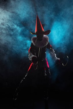 Kara cadılar bayramı teması için siyah ve kırmızı kostümlü soytarı ya da soytarı karakteri dramatik bir mavi duman ve siyah arka planda ışıklandırma ile sahnede duruyor..