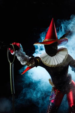 Çarpıcı bir soytarı ya da palyaço figürü canlı kırmızı ve siyah bir kostüm içinde, büyük bir konik şapka ve yaka, mavi dumanlı arka planda baloncuk asalar tutuyor. Cadılar Bayramı karakter teması.