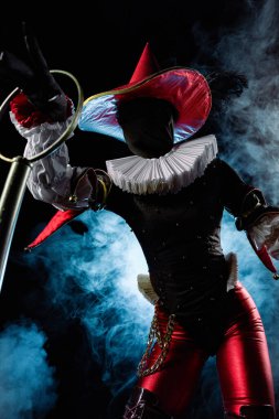 Çarpıcı bir soytarı ya da palyaço figürü canlı kırmızı ve siyah bir kostüm içinde, büyük bir konik şapka ve yaka, mavi dumanlı arka planda baloncuk asalar tutuyor. Cadılar Bayramı karakter teması.