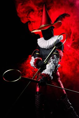 Striking mysterious jester or harlequin figure in vibrant red and black costume, with large conical hat with feather and ruff, holding bubble wands on red smoky background. Halloween character theme.