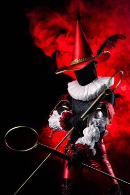 Striking mysterious jester or harlequin figure in vibrant red and black costume, with large conical hat with feather and ruff, holding bubble wands on red smoky background. Halloween character theme.