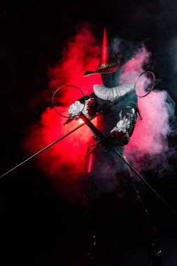 Striking mysterious jester or harlequin figure in vibrant red and black costume, with large conical hat with feather and ruff, holding bubble wands on red smoky background. Halloween character theme.