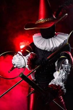 Kırmızı ve siyah kostümlü esrarengiz soytarı ya da palyaço figürü, tüy ve fırfırlı büyük konik şapkalı, kırmızı dumanlı arka planda baloncuk asaları tutan. Cadılar Bayramı karakter teması. Yakın plan..
