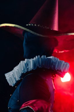 Dramatik yakın plan, esrarengiz soytarı ya da palyaço figürünün düşük açılı portresi konik şapkalı ve devasa beyaz yakalı koyu kırmızı ışık altında. Cadılar Bayramı karakter teması.