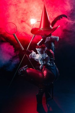 Kırmızı ve siyah kostümlü gizemli soytarı ya da palyaço figürünün dinamik ve dramatik çekimi kalın kırmızı duman ve ışığın arka planına şiddetle karşı duruyor. Cadılar Bayramı karakter teması.