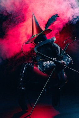 Kırmızı ve siyah kostümlü gizemli soytarı ya da palyaço figürünün dinamik ve dramatik çekimi kalın kırmızı duman ve ışığın arka planına şiddetle karşı duruyor. Cadılar Bayramı karakter teması.