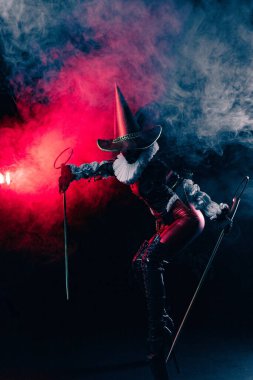 Kırmızı ve siyah kostümlü soytarı ya da palyaço kostümlü, elinde köpük değnekleri olan kırmızı ve mavi dumanla çarpıcı bir şekilde aydınlatılmış gizemli soytarının dinamik görüntüsü. Cadılar Bayramı karakter teması