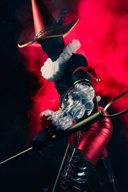 Detaylı siyah ve kırmızı bir kostüm içindeki gizemli soytarı ya da palyaço figürünün dinamik profil çekimi yoğun kırmızı dumanla çarpıcı bir şekilde aydınlatıldı. Cadılar Bayramı karakter teması.