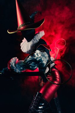 Detaylı siyah ve kırmızı bir kostüm içindeki gizemli soytarı ya da palyaço figürünün dinamik profil çekimi yoğun kırmızı dumanla çarpıcı bir şekilde aydınlatıldı. Cadılar Bayramı karakter teması.