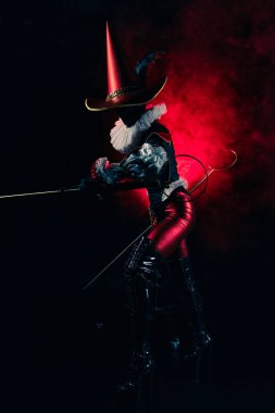 Detaylı siyah ve kırmızı bir kostüm içindeki gizemli soytarı ya da palyaço figürünün dinamik profil çekimi yoğun kırmızı dumanla çarpıcı bir şekilde aydınlatıldı. Cadılar Bayramı karakter teması.