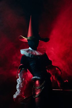 Dramatik soytarı kostümü ve şapkası giymiş bir sanatçının karanlık ve karamsar portresi. Kırmızı ve mavi aksanlı aydınlatma gizemli, gotik ve ürkütücü bir atmosfer yaratır. Cadılar Bayramı karakter teması.