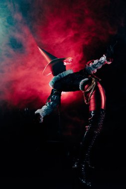 Dramatik soytarı kostümlü bir sanatçının dinamik ve esnek duruşu. Canlı kırmızı dumanlı ve cesur sanatsal ışıklandırmalı karanlık tiyatro stüdyosu. Cadılar Bayramı karakter teması.