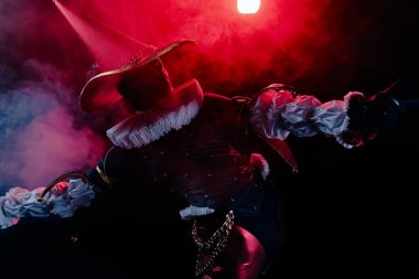 Özenle hazırlanmış fırfırları ve altın zinciri olan koyu renk kostümlü tiyatrocu. Canlı kırmızı dramatik ışık ve dumanla aydınlatılmış dinamik bir poz. Cadılar Bayramı karakter teması.