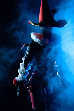 Kalın mavi dumana gizlenmiş gizemli bir soytarının ürkütücü ve atmosferik portresi. Dramatik ışıklandırma kostüm detaylarını ve uzun şapkayı ön plana çıkarıyor. Cadılar Bayramı karakter teması.