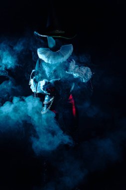 Yoğun mavi dumanın içinde duran gizemli soytarı karakterinin karanlık ve dramatik görüntüsü. Yoğun ışıklandırma, beyaz yaka ve uzun şapka. Cadılar Bayramı karakter teması.